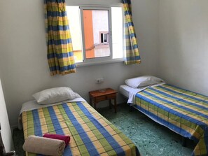 Une petite pièce avec deux lits, une fenêtre avec des rideaux, une table de chevet et une prise électrique au mur.