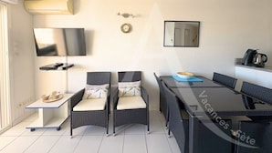 A compact living space with a sofa, two chairs, a dining table, a TV, and a small kitchen area.