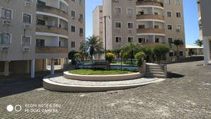 Un moderno edificio de apartamentos con varios balcones, un área ajardinada central con una fuente y una calle adoquinada.