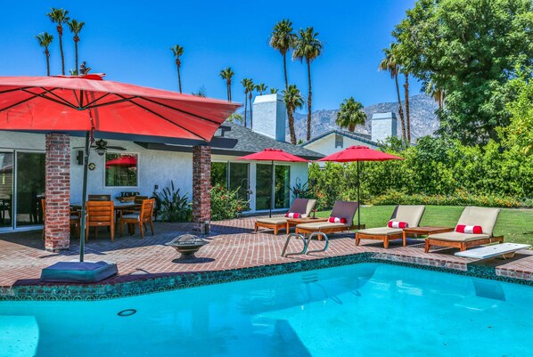 A pool area with lounge chairs and umbrellas, surrounded by palm trees and a brick patio.