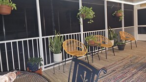 A covered outdoor area with a wooden floor, a small round table, and several wicker chairs. There are potted plants and a dog visible. - Steinhatchee Nest House (Steinhatchee)