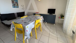 Ein Essbereich mit Tisch, bedeckt mit einer gemusterten Tischdecke, gelbe Stühle, eine Couch, ein Fernseher und ein Schrank.