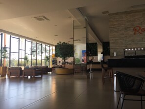 Een ruime lobby met grote ramen, een receptiebalie en een zithoek met stoelen en een plant.