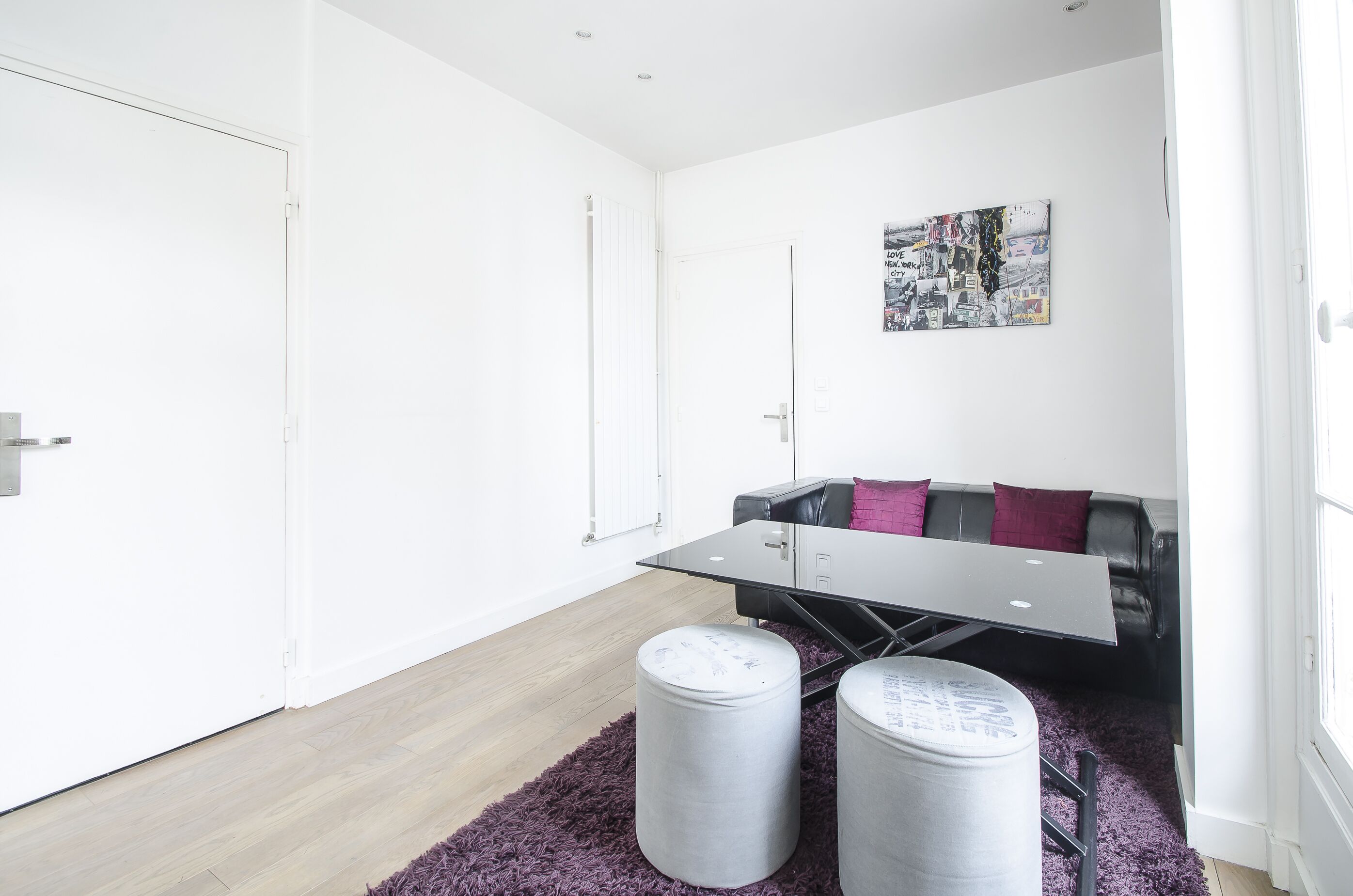 A modern living room with a glass-top table, a black leather sofa with red cushions, and a purple rug.