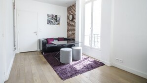 Uma sala de estar moderna com um sofá de couro preto, uma mesa de centro de vidro e dois pufes cinzas sobre um tapete roxo. Uma parede de tijolos e uma janela grande proporcionam iluminação natural.