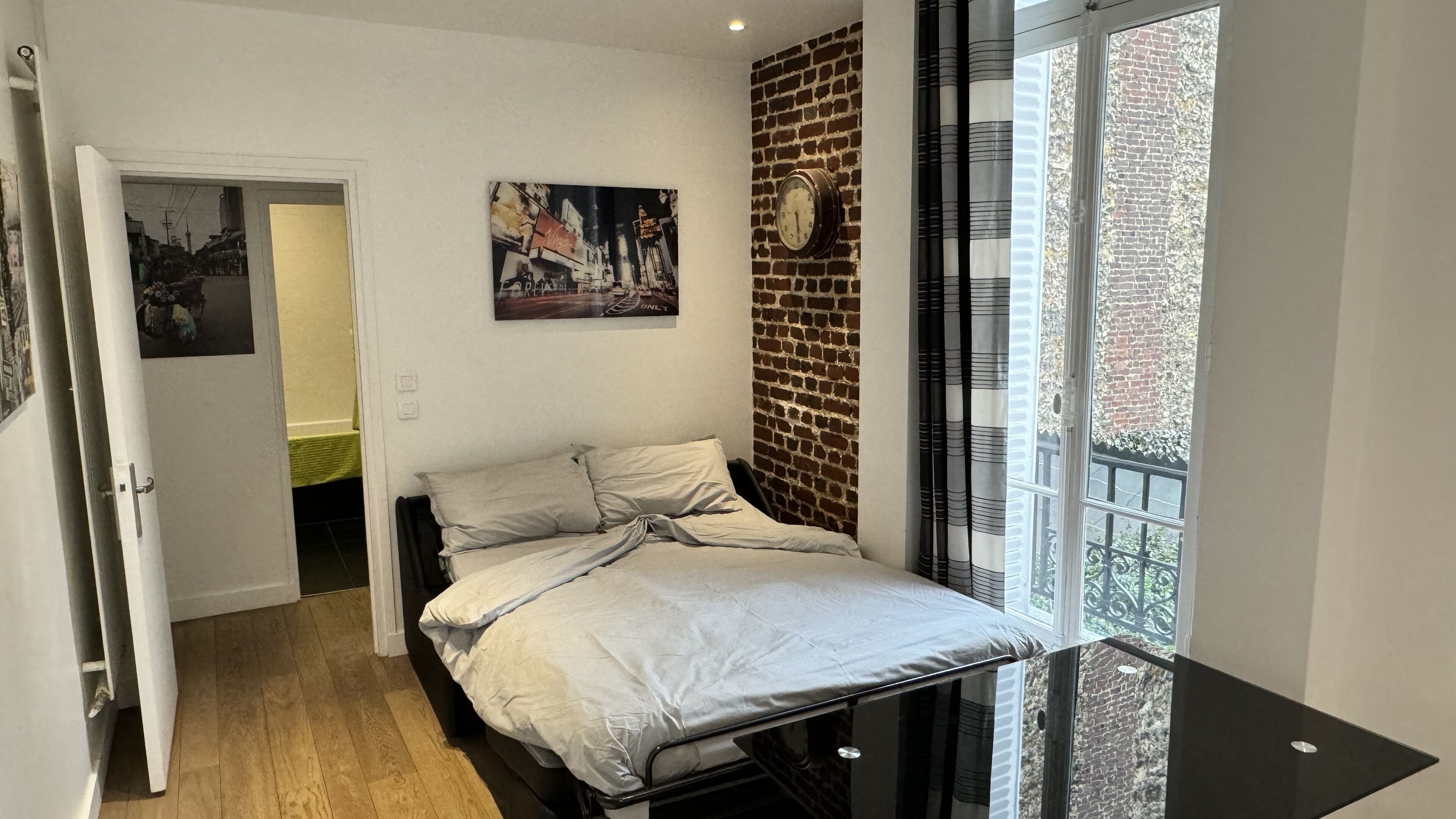 Une chambre à coucher avec un lit, une horloge au mur, une porte et une fenêtre donnant sur un mur de briques.