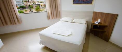 A hotel room with a bed, a nightstand, a painting, and a view of the city through the window.