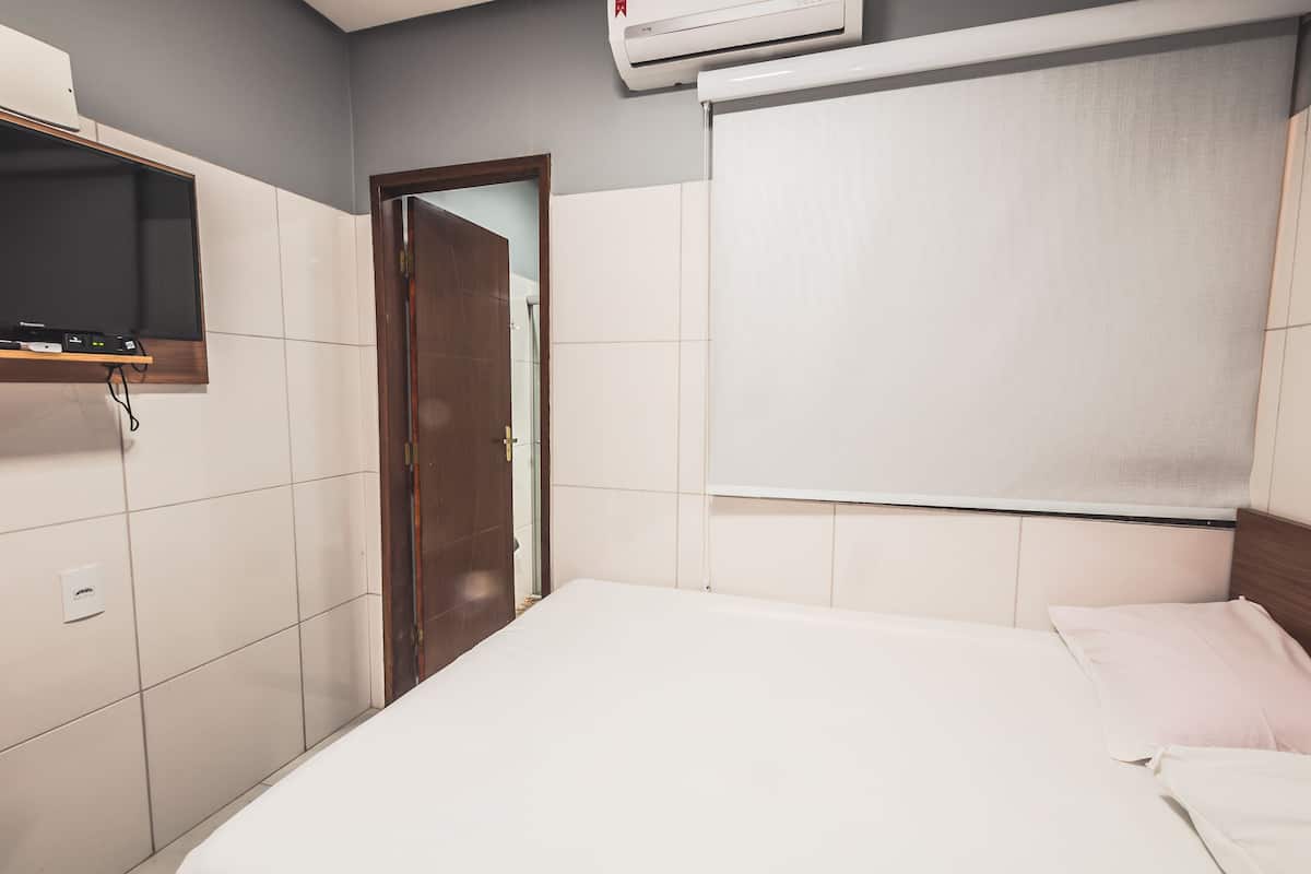 A hotel room with a bed, a television mounted on the wall, and a window with a blind.