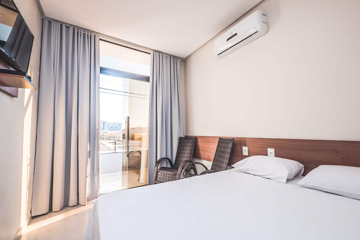 A hotel room with a bed, two chairs, a TV, and an air conditioning unit.