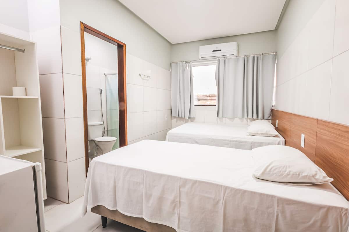 A hotel room with two beds, a shower, a toilet, and a window with curtains.