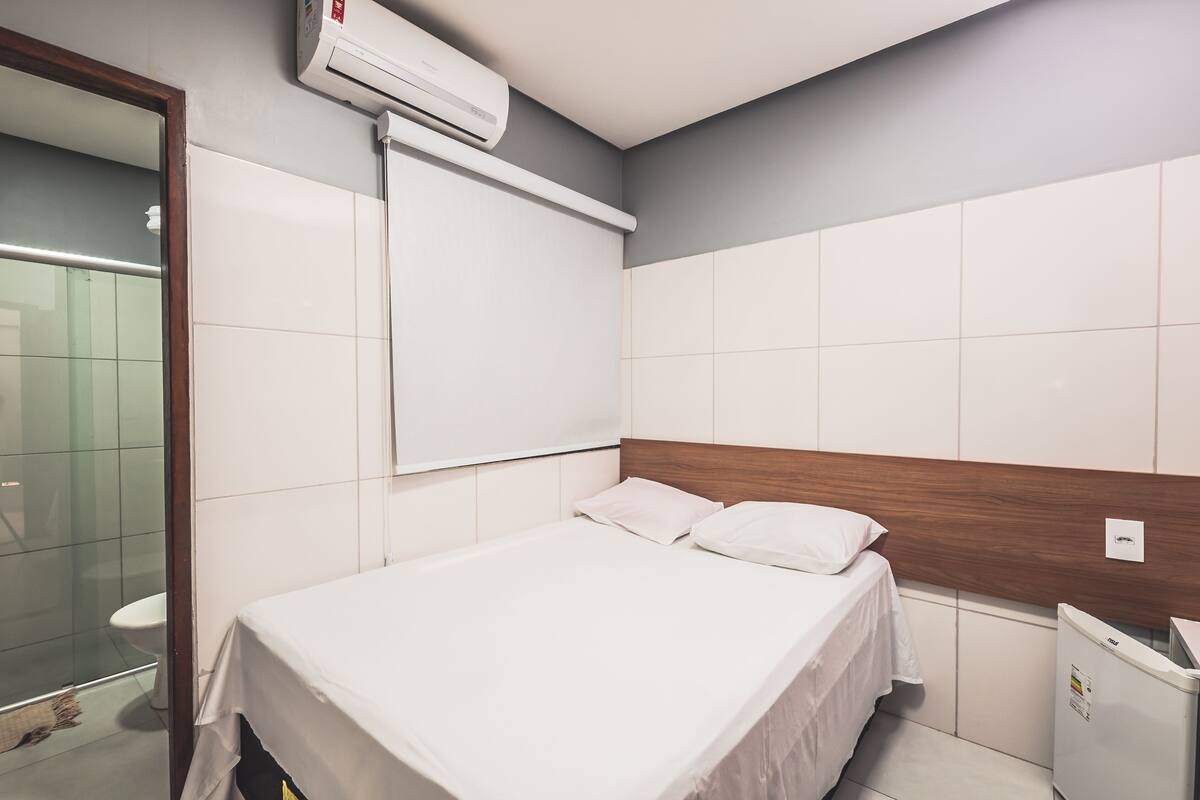 A hotel room with a bed, a wooden headboard, a white air conditioning unit, a white trash bin, and a mirror.