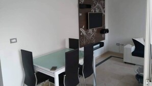 Habitación compacta con una mesa de comedor, dos sillas, un sofá pequeño blanco y un panel decorativo con un televisor incorporado.