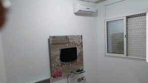 Una habitación con televisor de pared, aire acondicionado, ventana con persianas y un tapete rosa en el suelo.