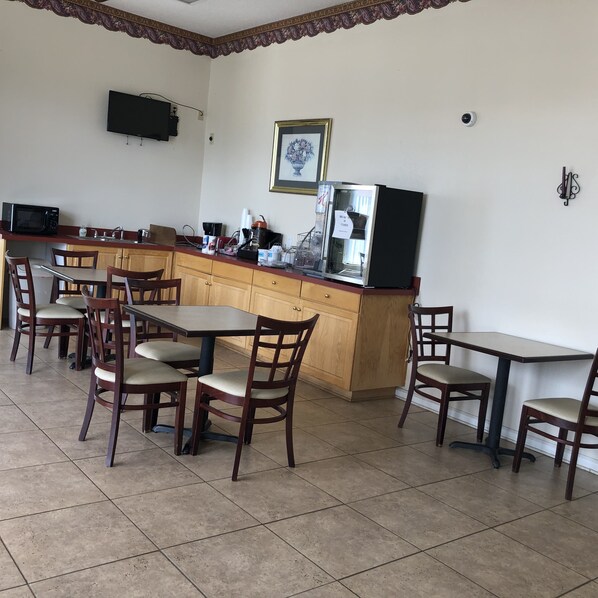 A small, clean kitchen with a microwave, coffee maker, and a refrigerator. There are wooden chairs and tables arranged around the kitchen.