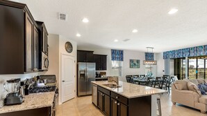 A modern kitchen with dark cabinetry, a large island, and a dining area with a view. - Solterra Sunshine - 5* - South facing large pool/spa - game room, gated resort (Davenport)