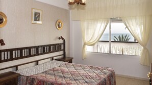 A bedroom with a bed, a wooden headboard, a nightstand, a bedside table, a window with curtains, and a view of the sea. - LIDER, 4-D (Cullera)