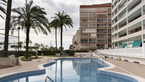 A swimming pool with a clear blue water, surrounded by a white fence, palm trees, and a multi-story building with balconies. - LIDER, 4-D (Cullera)