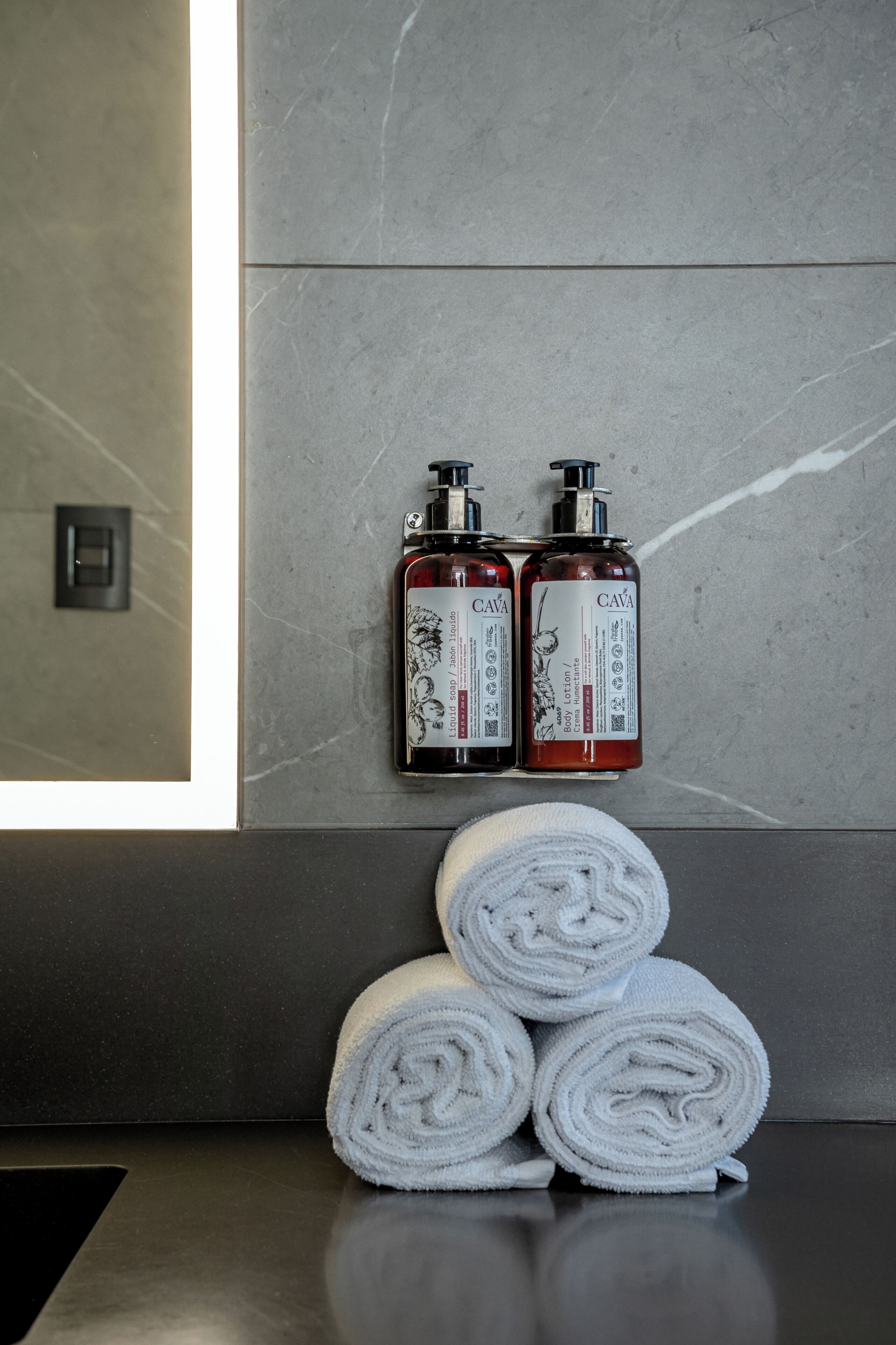 A bathroom with a marble wall, two CAVA body lotion bottles, and three rolled towels on a counter.