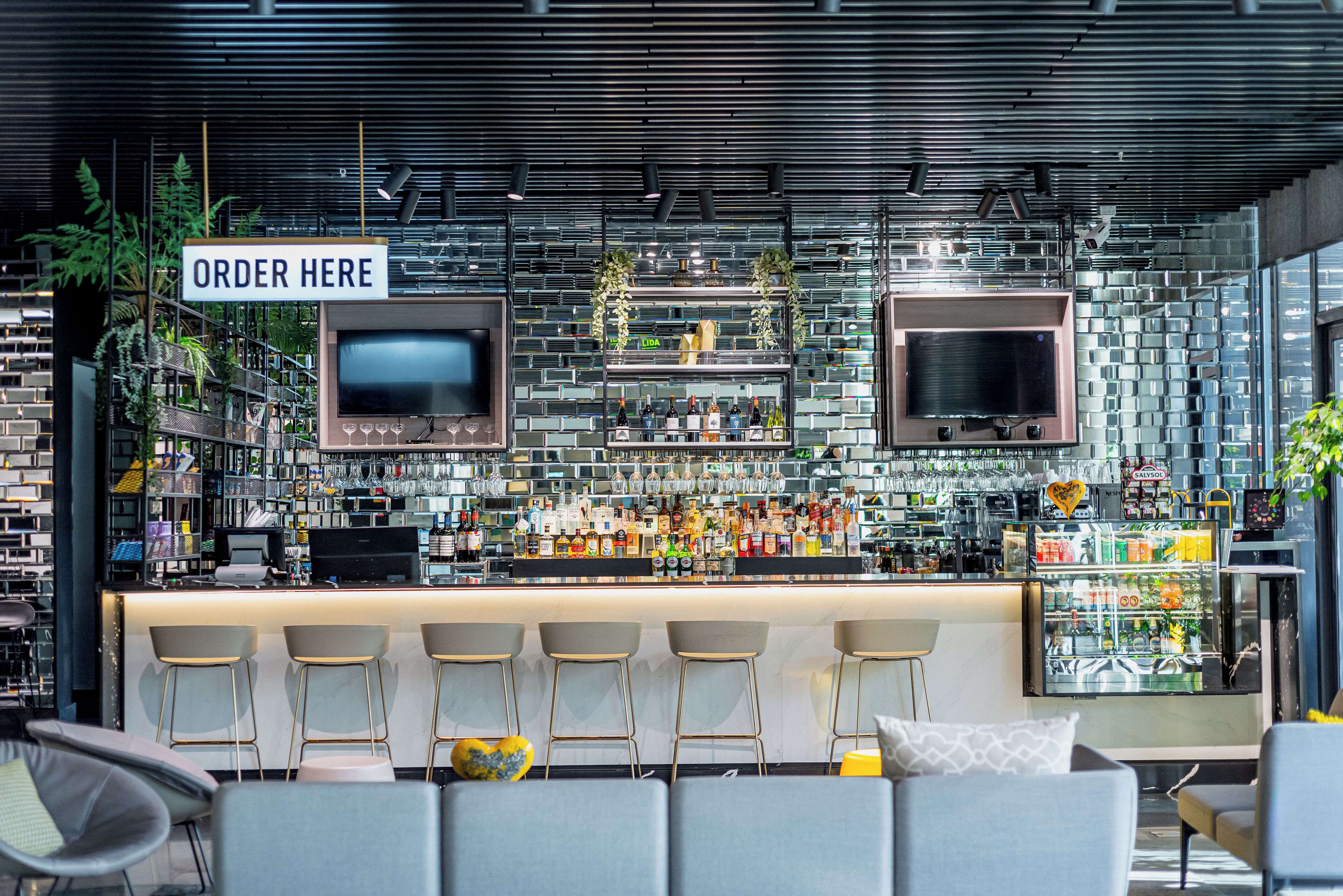 Un área de bar moderna con un letrero que dice 'ORDER HERE', taburetes y una variedad de botellas y vasos en estantes.