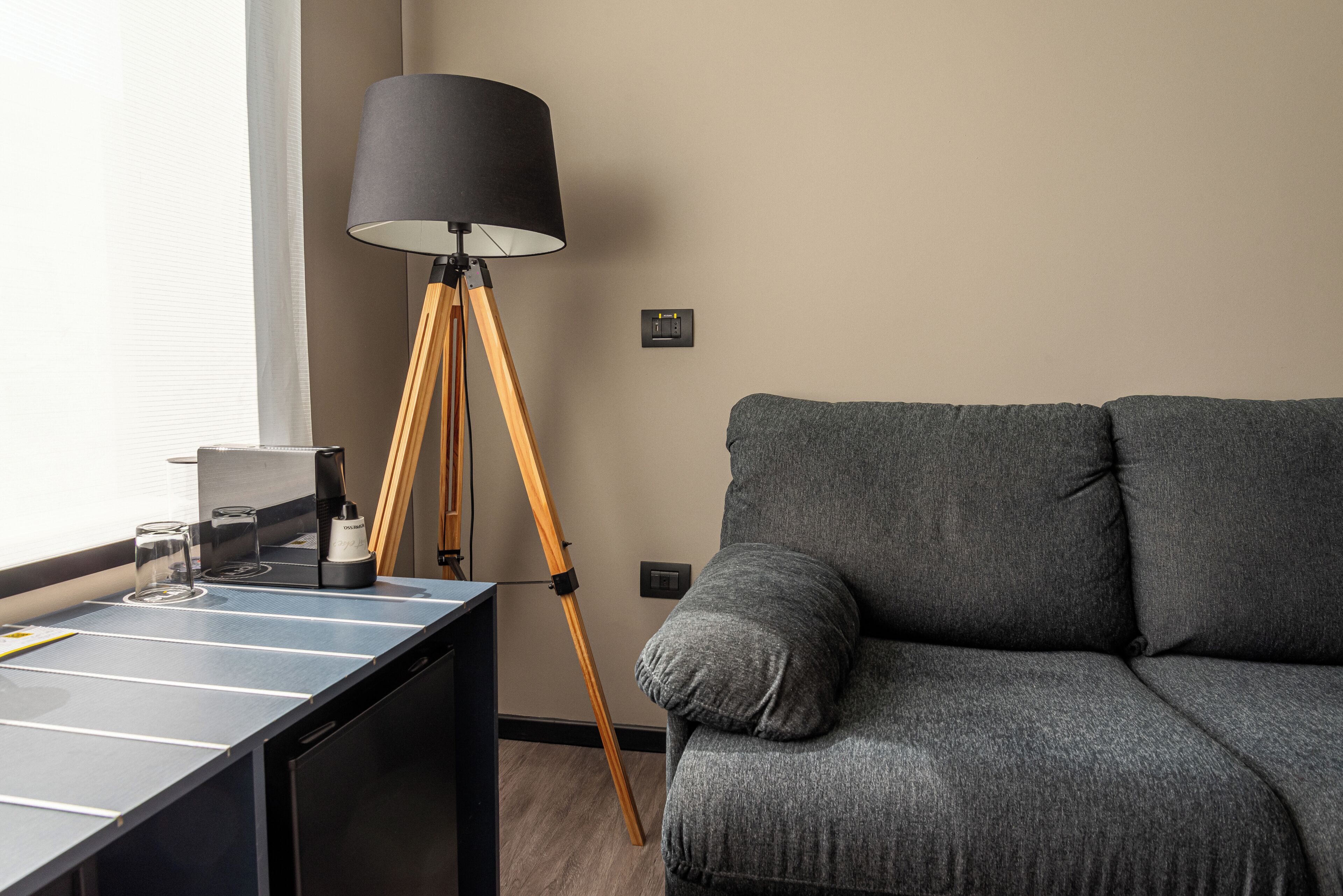 Una sala moderna con un sofá gris, una lámpara de pie con pantalla negra, una mesita con una cafetera y un enchufe en la pared.
