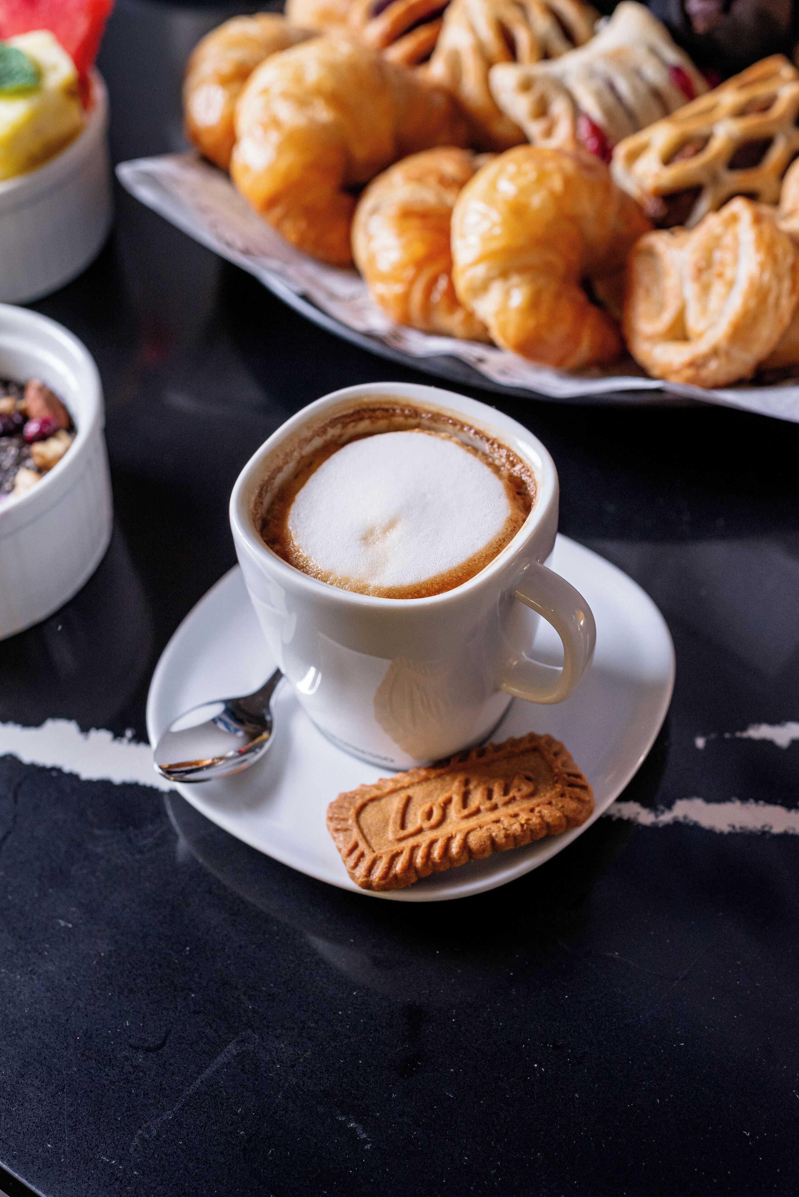 Una taza de café con una cuchara en un plato, una galleta y un plato de pasteles sobre una superficie oscura.