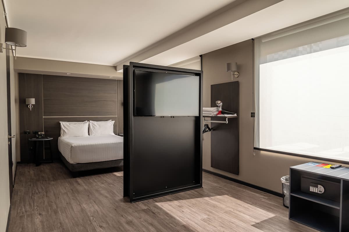 A modern hotel room with a bed, a desk, a wardrobe, and a window.