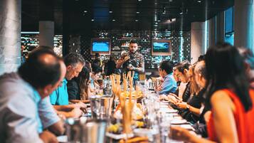 Een restaurant met een lange, gedeelde tafel, meerdere schermen aan de muur en een bartender die drankjes uitserveert.