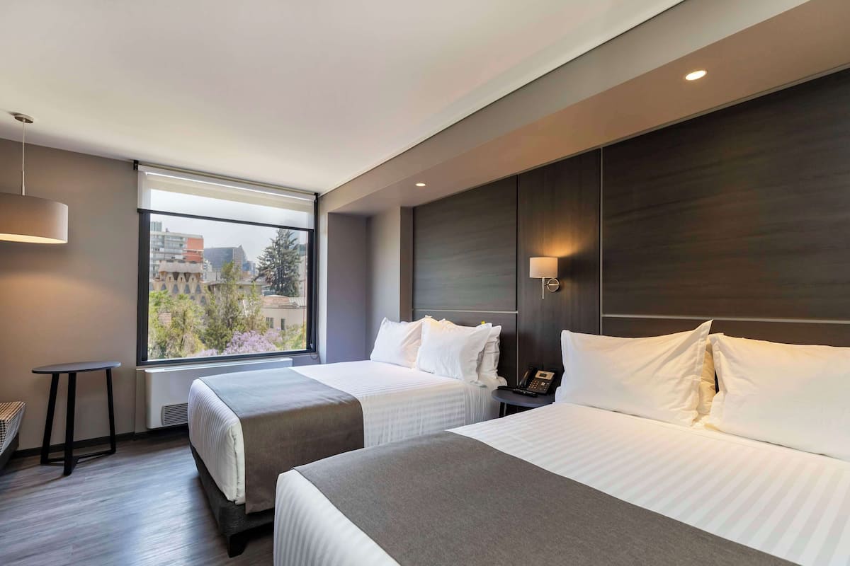 A modern hotel room with two beds, a small table, a lamp, and a view of the city.
