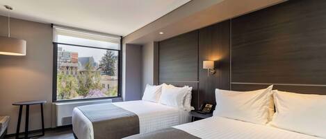 A modern hotel room with two beds, a small table, a lamp, and a view of the city.