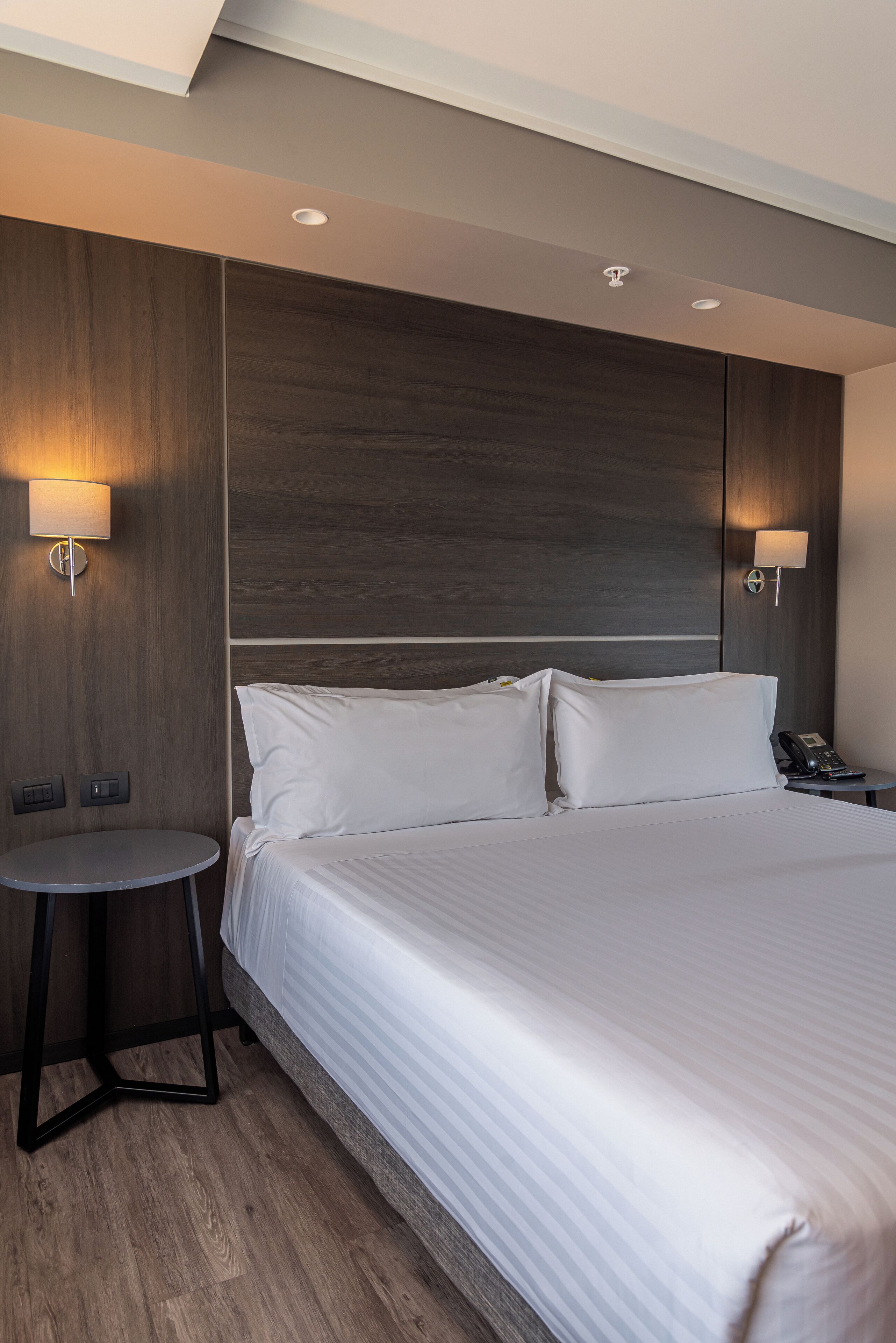A hotel room with a large bed, two bedside lamps, a small round table, and a telephone on the nightstand.
