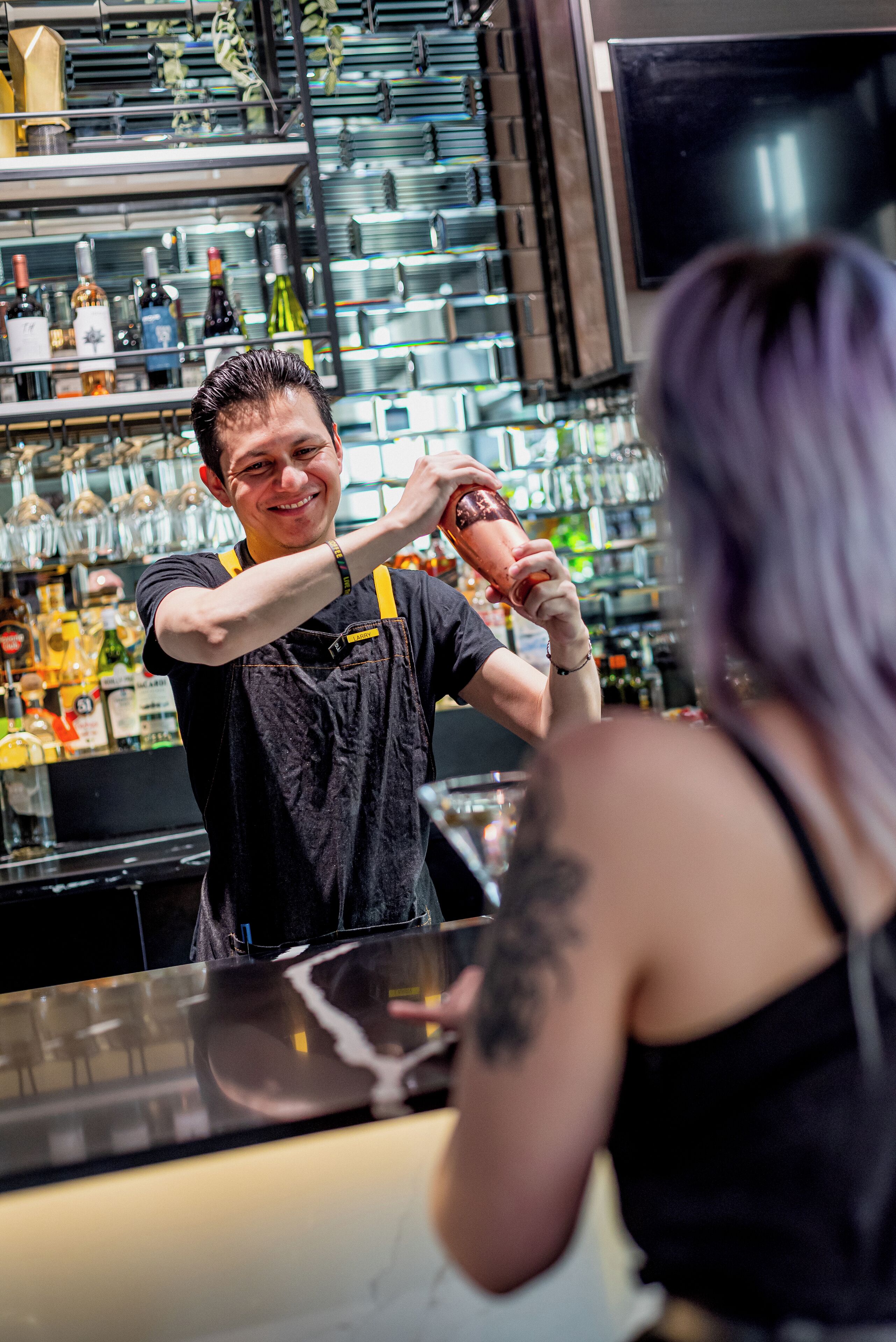 Un cantinero preparando una bebida detrás de una barra con estantes llenos de botellas y vasos.