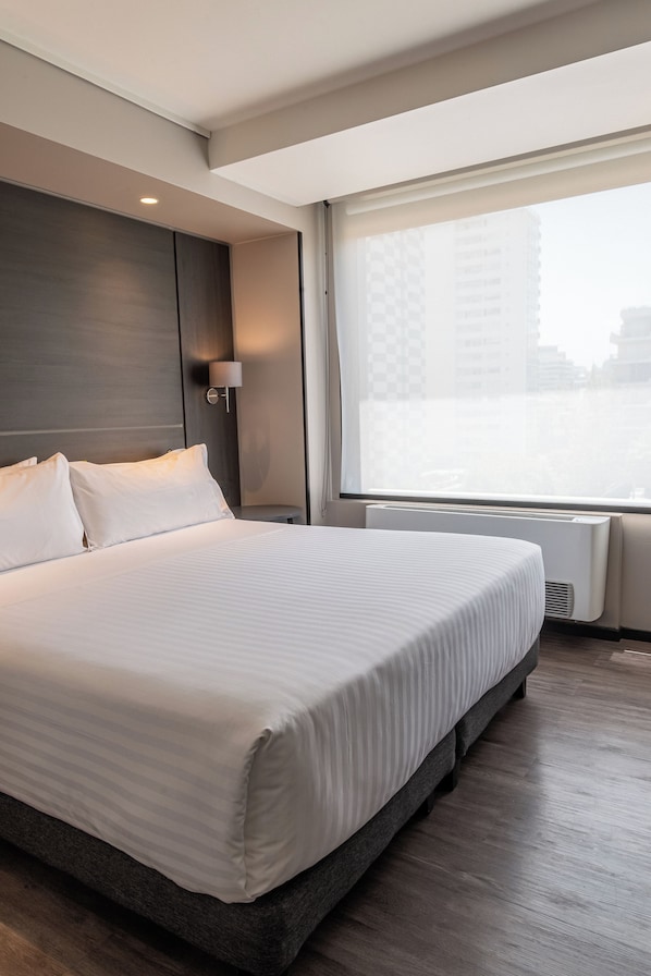 Une chambre d’hôtel moderne dotée d’un grand lit, d’une lampe de chevet, d’une fenêtre donnant sur des immeubles et d’un climatiseur fixé au mur.