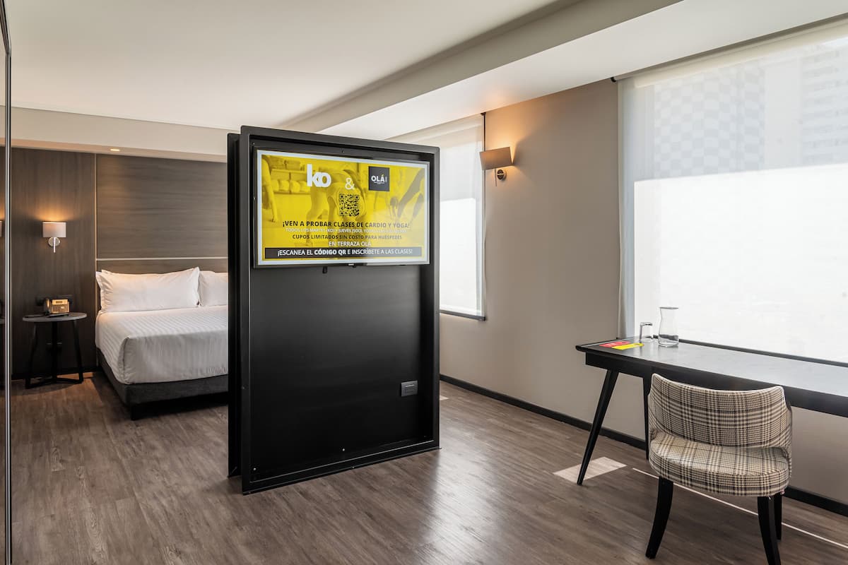 A hotel room with a bed, a desk, a chair, a TV screen with a QR code, and a window with a view.