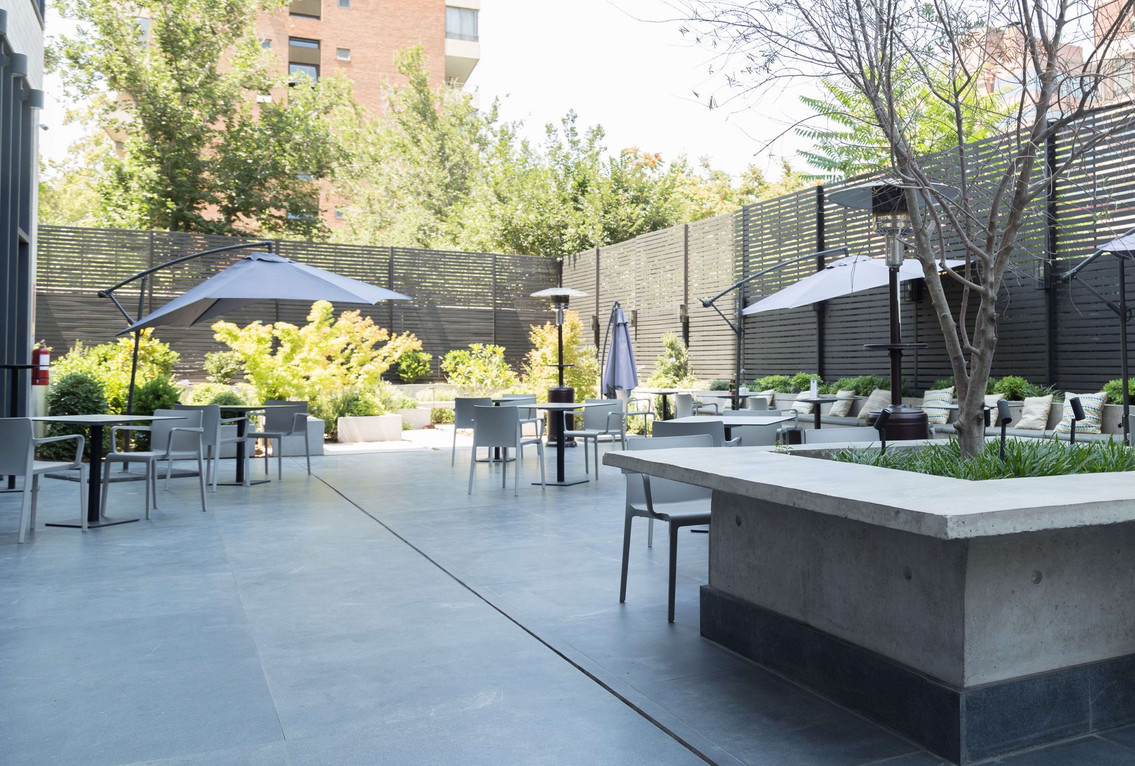 Un patio al aire libre con mesas, sillas, sombrillas y una jardinera de concreto con plantas.