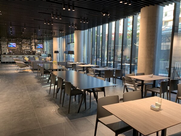 Un comedor moderno con mesas largas y sillas, ventanas amplias y una zona de bar con televisores.