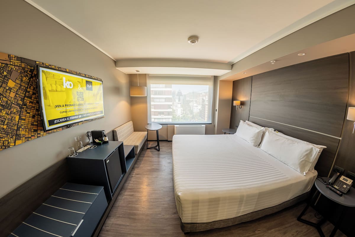 A modern hotel room with a large bed, a small desk, a sofa, a TV mounted on the wall, and a window with a city view.