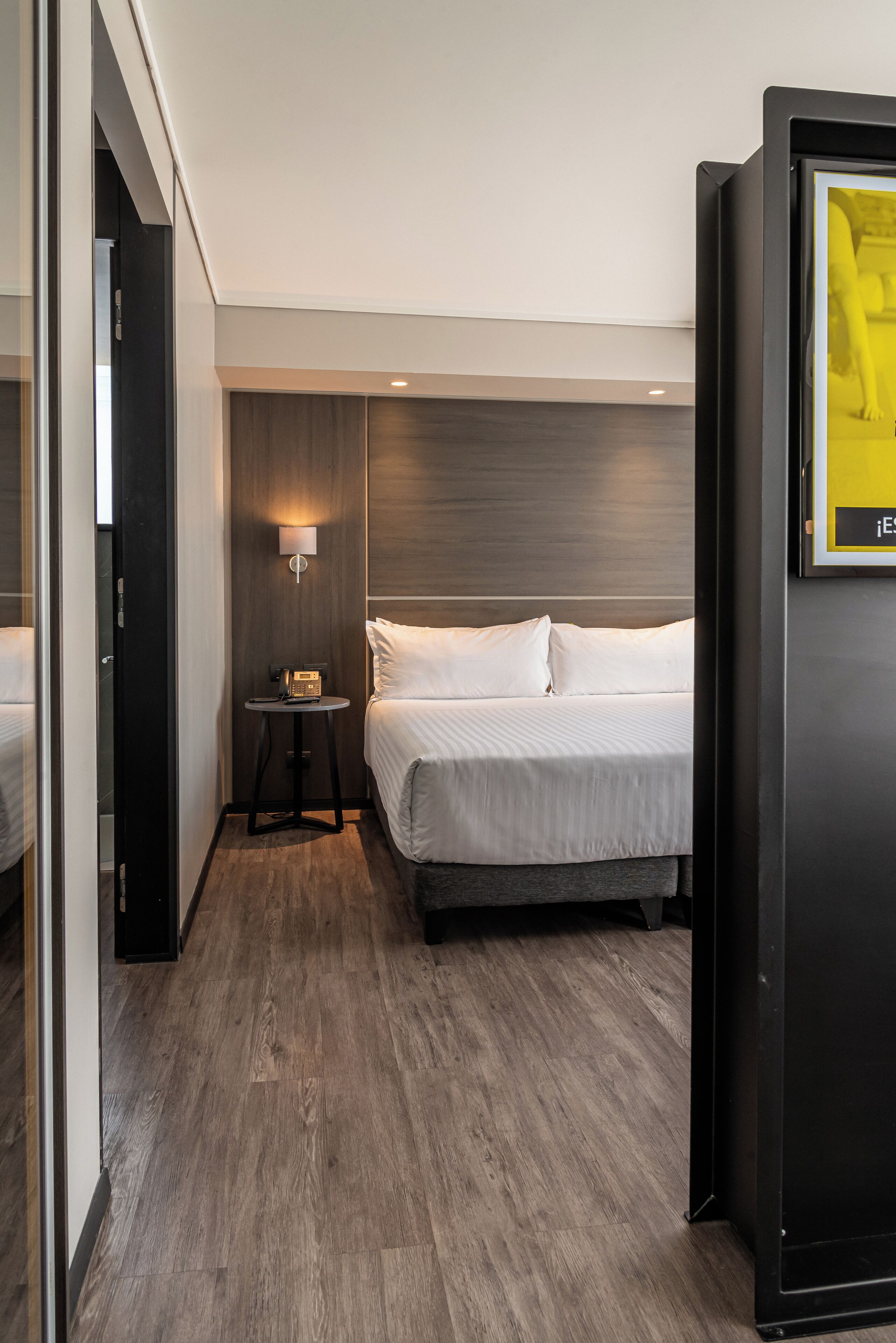 A hotel room with a bed, bedside table, lamp, and a framed picture on the wall.