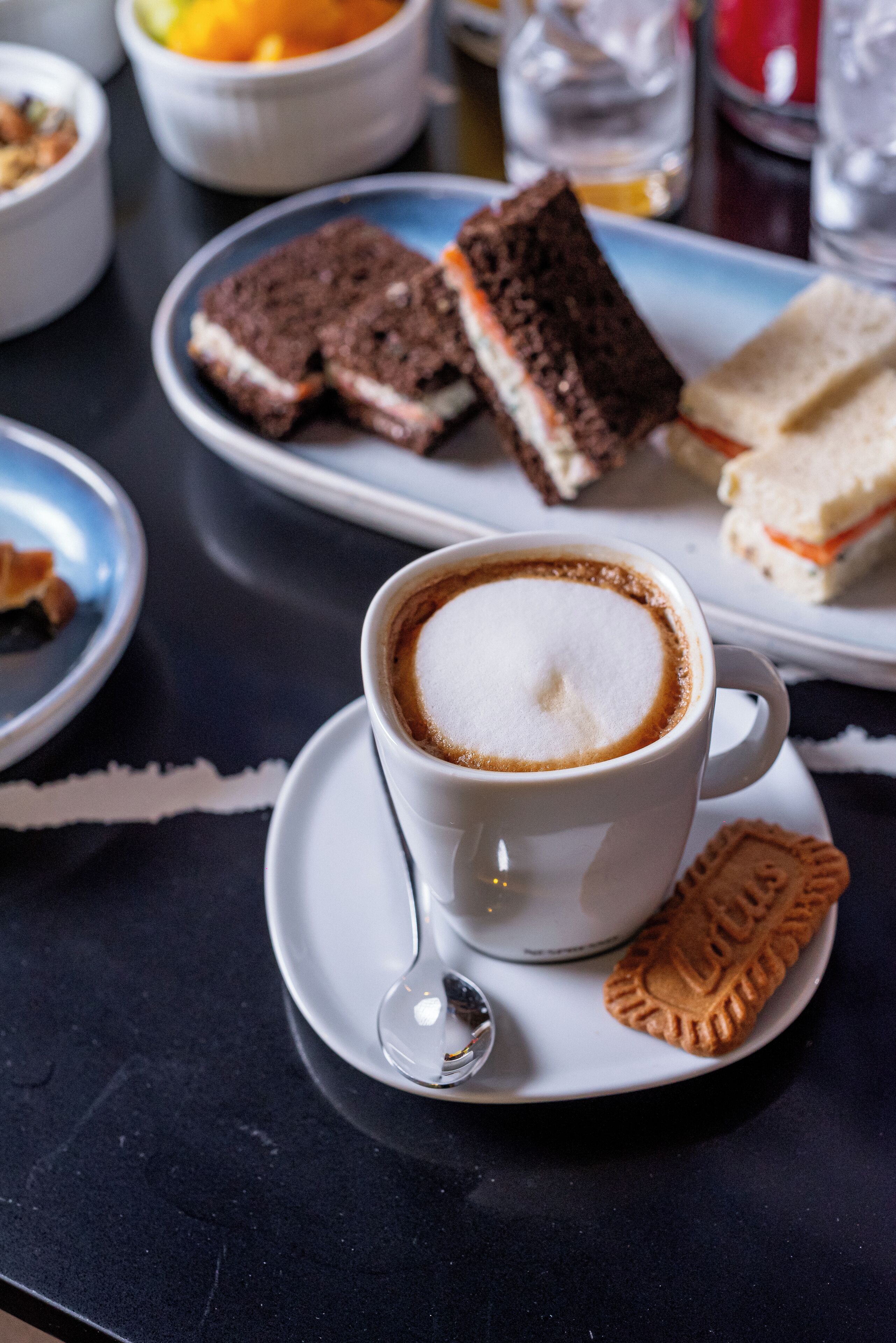 Una taza de café con espuma, sobre un platito, acompañada de una galleta y un sándwich en un plato.