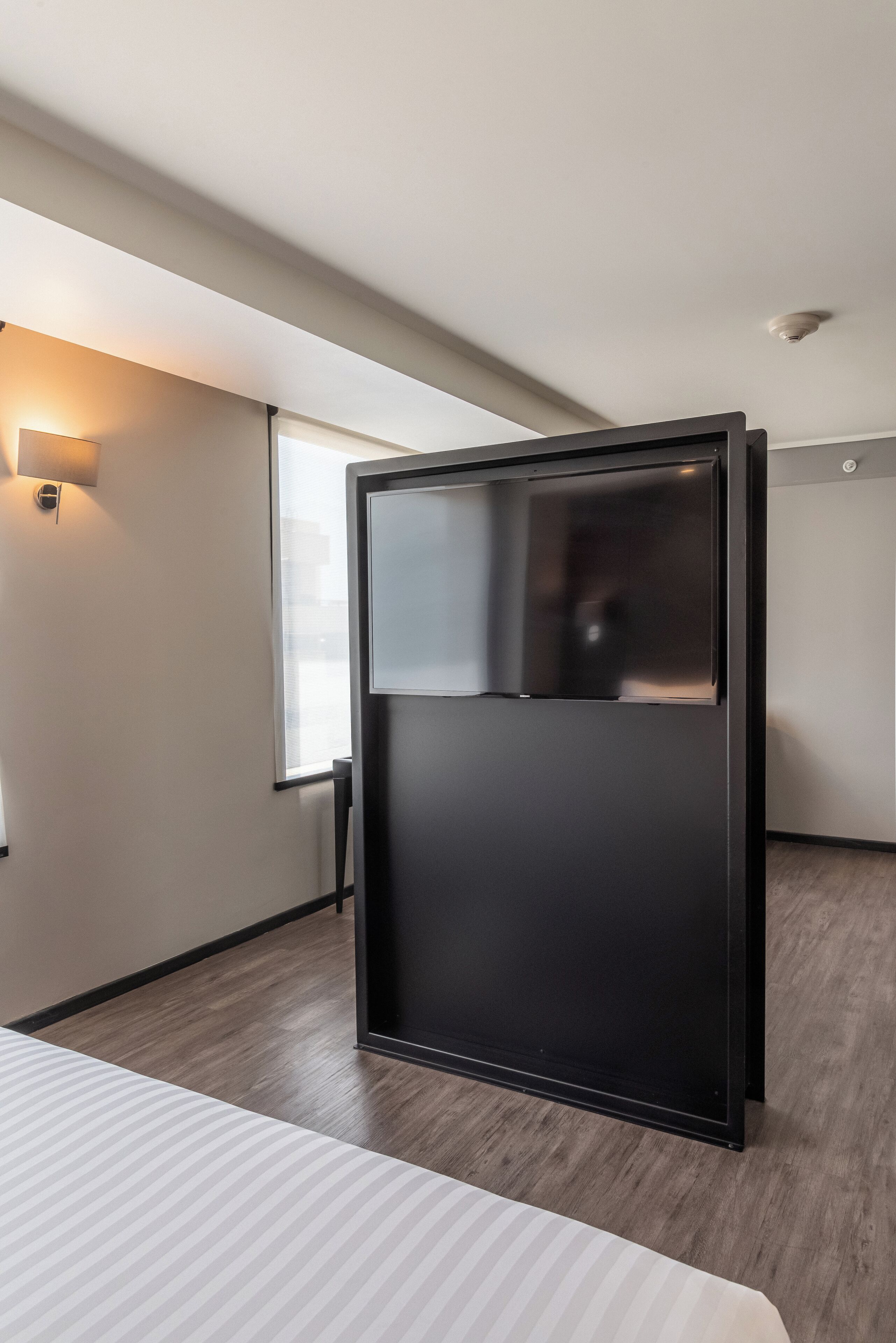 A modern hotel room with a large window, a bed with striped bedding, and a black wardrobe.