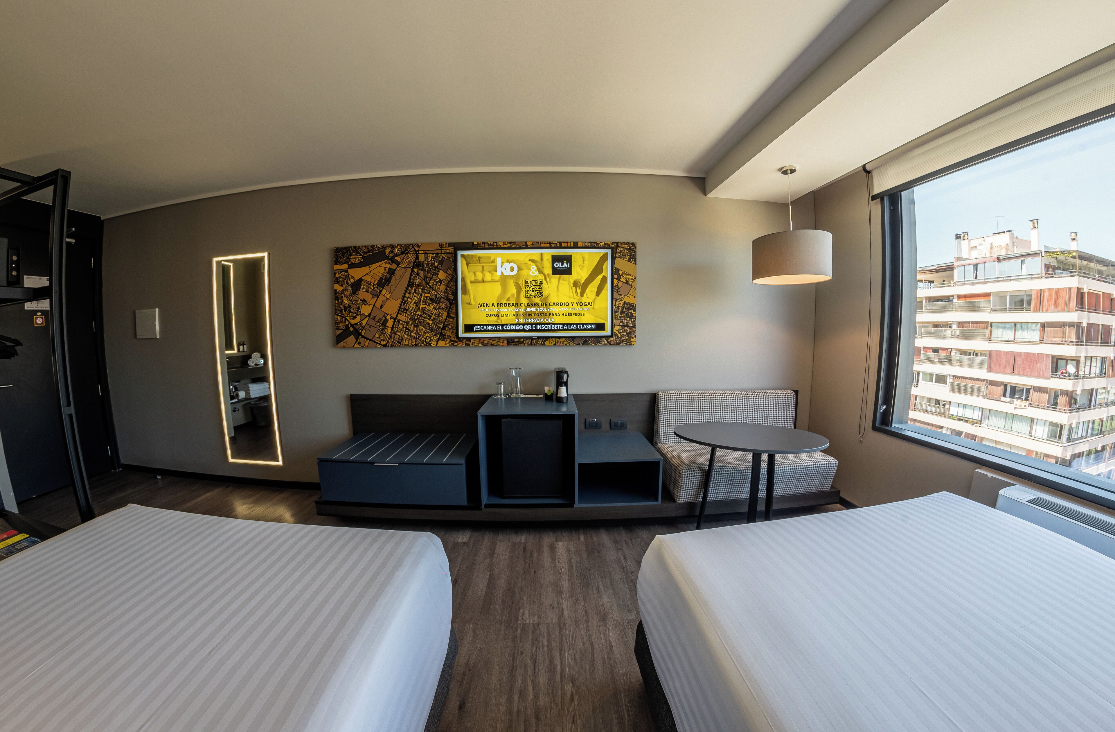 Habitación de hotel con dos camas, un sofá, una mesita, un televisor y ventana con vistas a la ciudad.