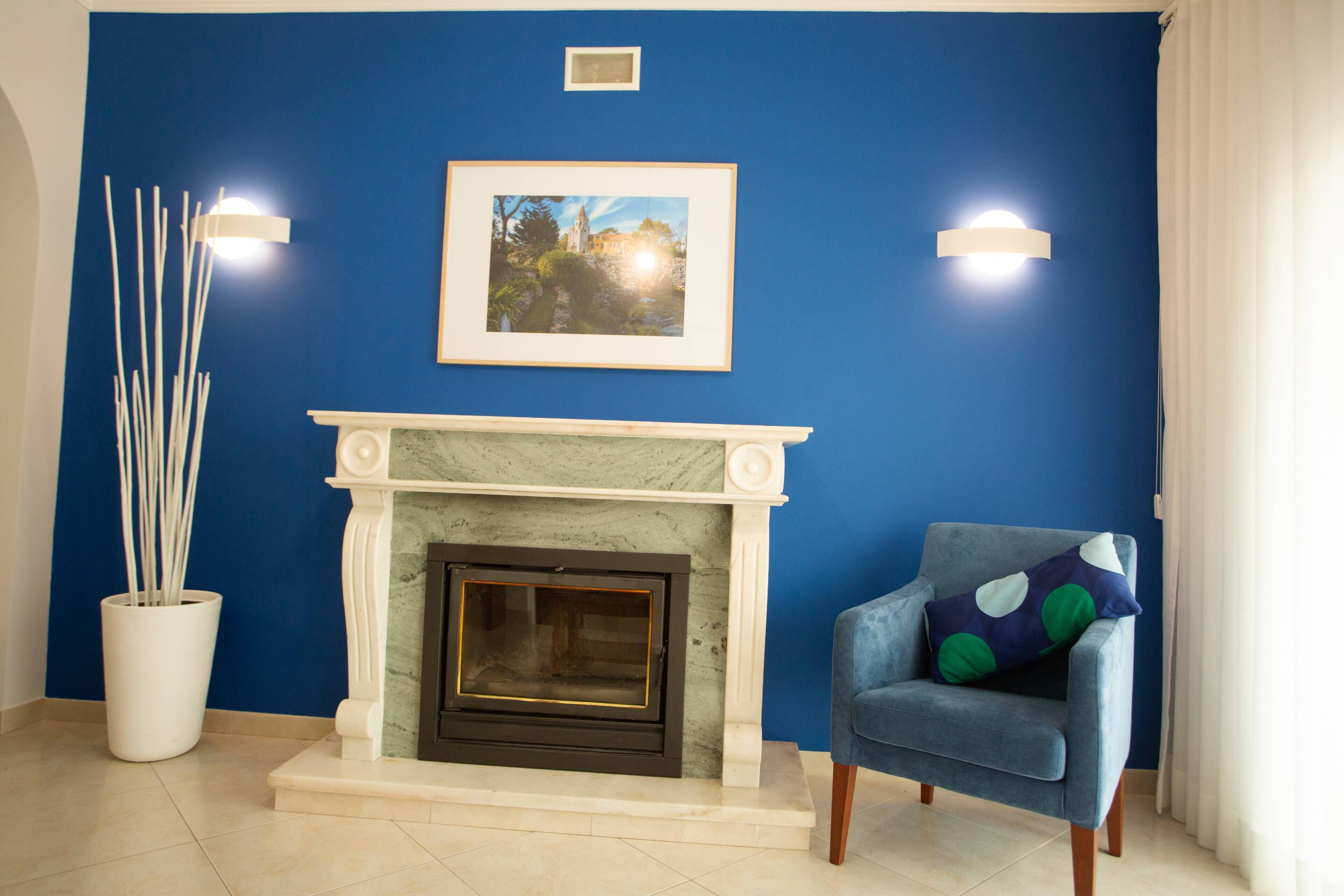 Une pièce avec un mur bleu, une cheminée ornée d’un manteau en marbre, un tableau encadré au-dessus de la cheminée, un fauteuil bleu avec un coussin à pois et deux appliques murales.