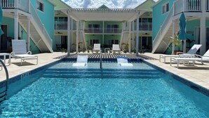 A swimming pool with lounge chairs and a covered seating area, surrounded by multi-story buildings with balconies. - The No Problem Ocean View Studio Cottage in East End (East End)