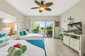 Habitación de hotel con dos camas, ventilador de techo, televisor y balcón con vista a palmeras.