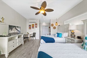 A hotel room with two beds, a desk with a chair, a ceiling fan, and a TV.