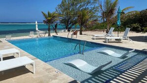 Una piscina con agua cristalina azul, rodeada de sillas de descanso y sombrillas blancas, con vistas al océano y palmeras.
