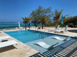 A pool with clear blue water, surrounded by white lounge chairs and umbrellas, with a view of the ocean and palm trees.
