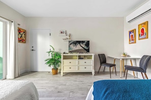A room with a bed, a white dresser, a desk with two chairs, a potted plant, and artwork on the walls.