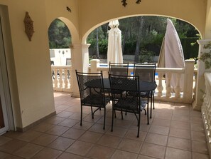 Une terrasse couverte avec une table et des chaises, un parasol blanc et une vue sur une piscine et la verdure.