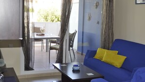 Un salon avec un canapé bleu, une petite table et un fauteuil. Une porte vitrée donne sur un espace extérieur aménagé avec une table et des chaises.