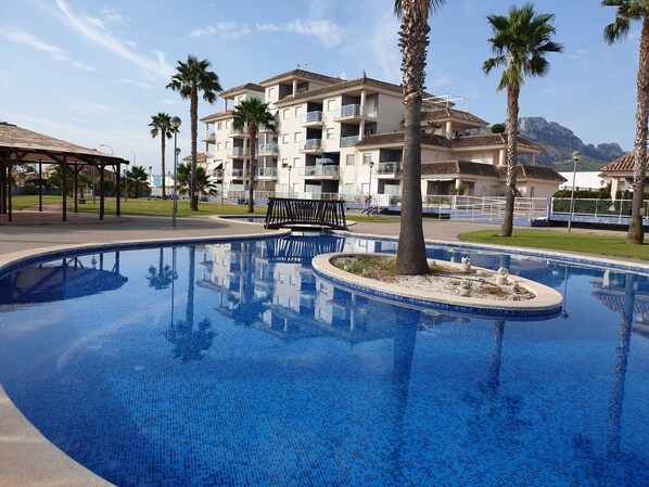 A swimming pool with tiled blue mosaic, surrounded by palm trees and a building with balconies.